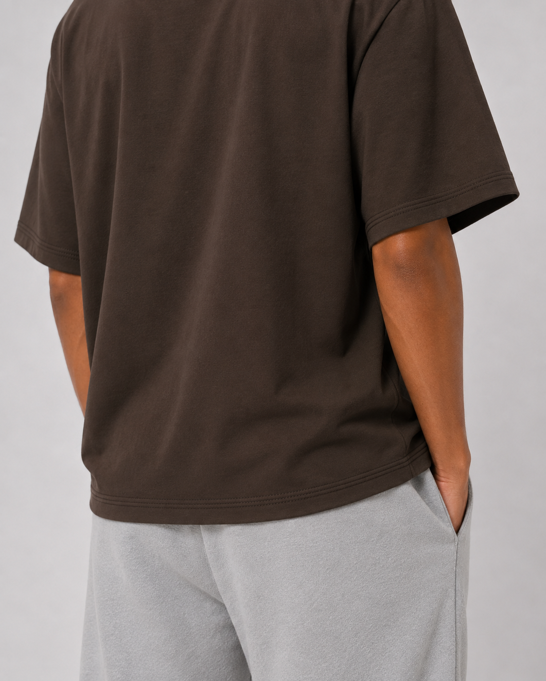 A person wearing an oversized dark brown short-sleeve t-shirt and light gray sweatpants, shown from the back against a plain light gray studio background.