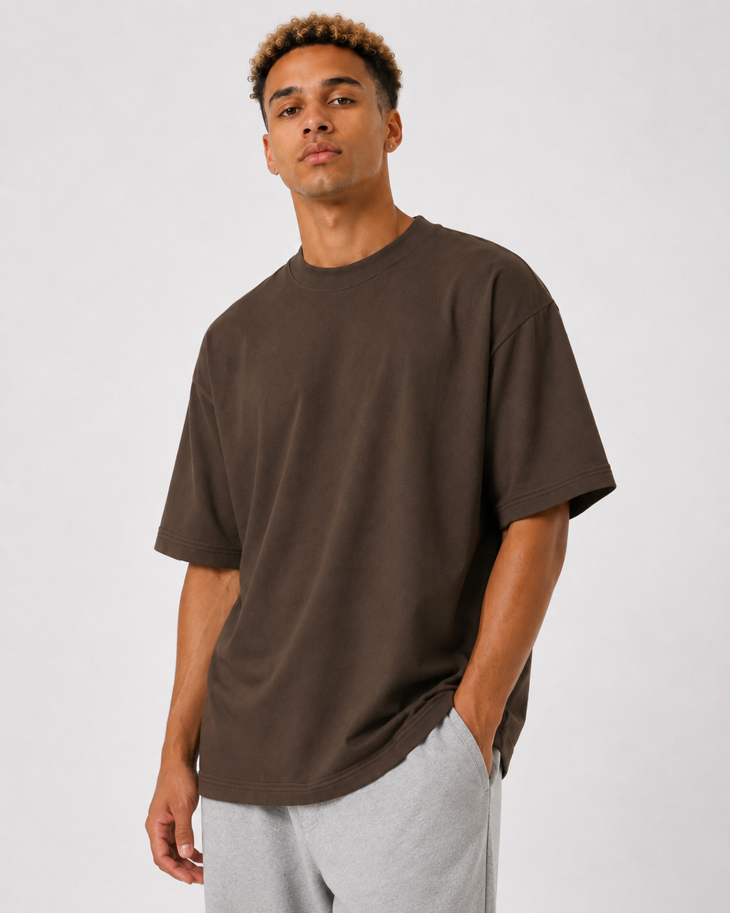 A man wearing an oversized dark brown crewneck t-shirt and light gray sweatpants, standing against a light gray studio background.