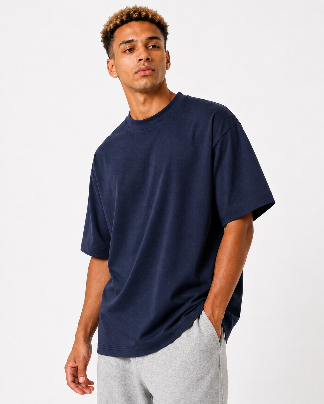 A man wearing an oversized navy blue crewneck t-shirt and light gray sweatpants, standing against a plain white studio background.