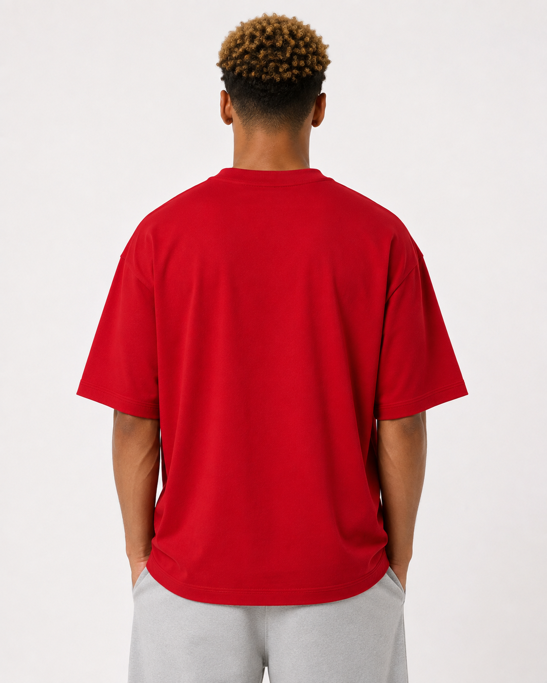 A man viewed from behind wearing a bright red oversized short-sleeve t-shirt and light gray sweatpants, standing against a plain white studio background.