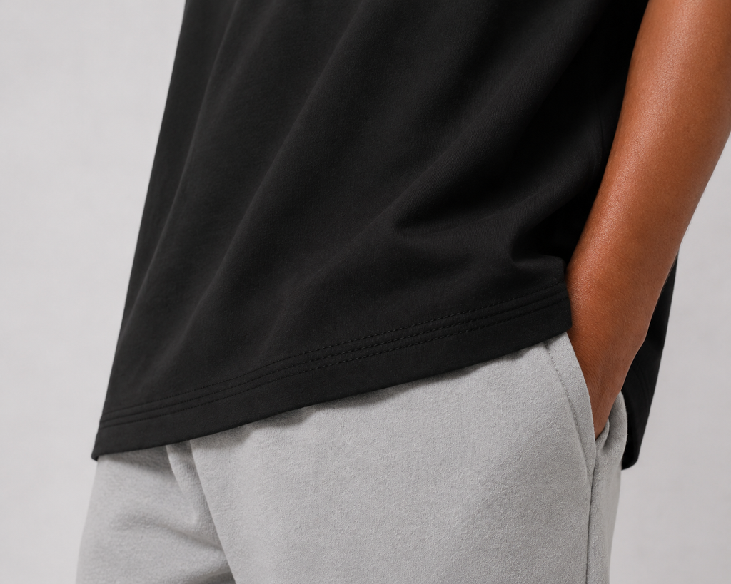 Close-up of a person wearing a black short-sleeve top and light gray sweatpants, with the black shirt hem and side seam visible against a plain light gray background.