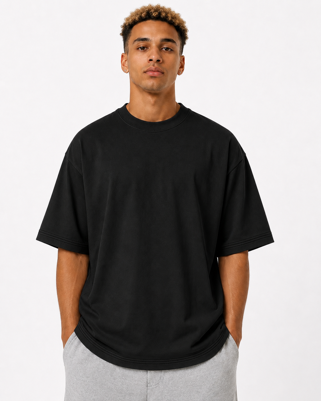 A man wearing an oversized black short-sleeve crewneck t-shirt and light gray sweatpants, standing against a plain white studio background.