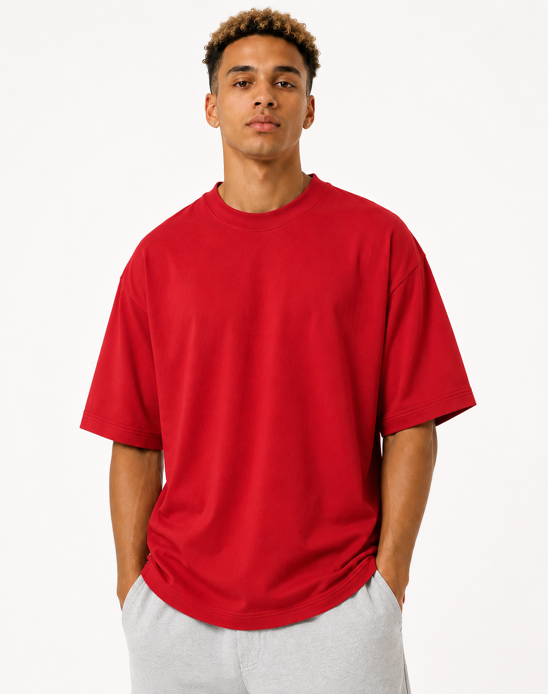 A man wearing an oversized bright red short-sleeve crewneck t-shirt and light gray sweatpants, standing against a plain white studio background.